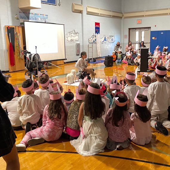 Children wearing Piglet Mindset headbands sitting in a school gymnasium, watching Melissa with Piglet, Zoey, and Evie.